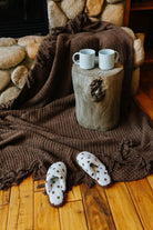 Brown waffle fringe blanket draped beside a fireplace with blue mugs and polka dot slippers.