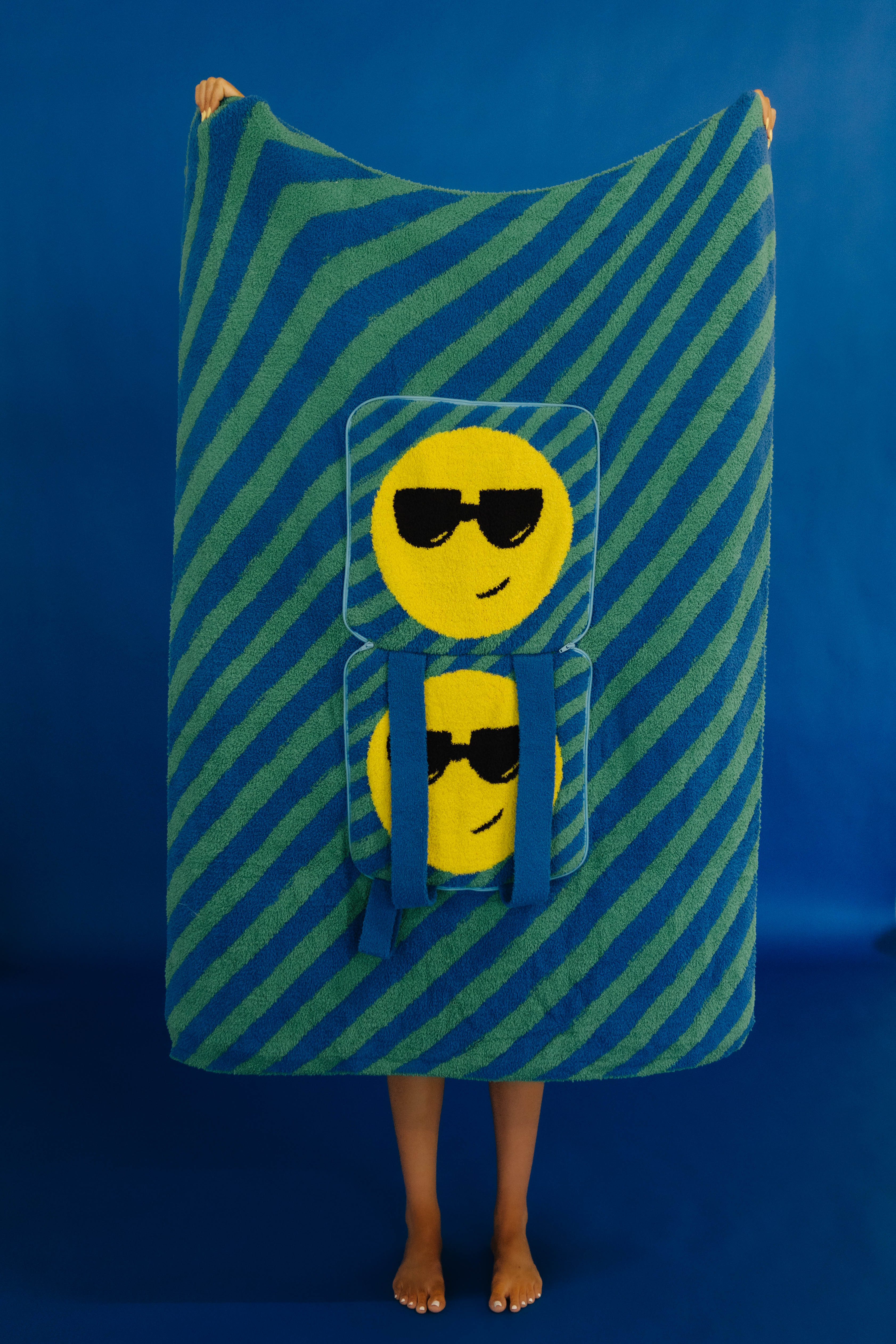A person standing barefoot against a blue studio backdrop, holding up a large rectangular quillow with diagonal blue and green stripes and two yellow emoji faces wearing sunglasses printed on the front.