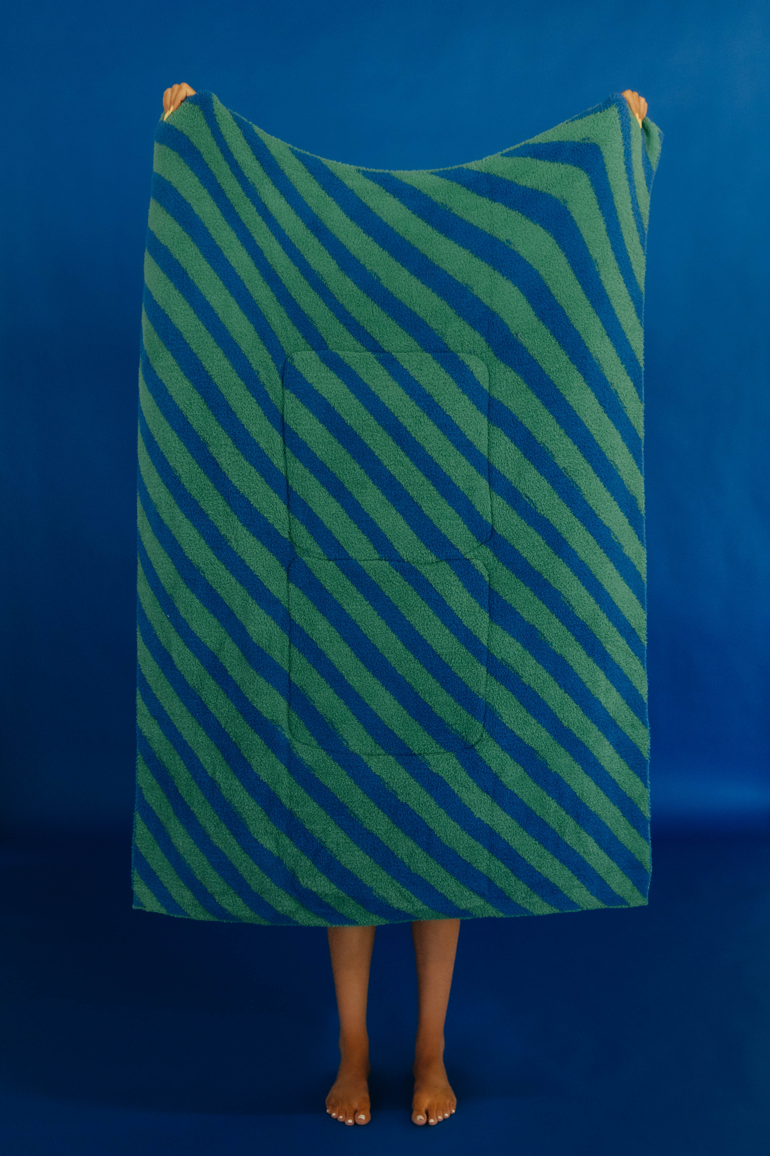 A person standing barefoot against a solid blue backdrop, holding up a large rectangular quillow with diagonal green and blue stripes and a central pocket.