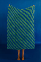 A person standing barefoot against a solid blue backdrop, holding up a large rectangular quillow with diagonal green and blue stripes and a central pocket.