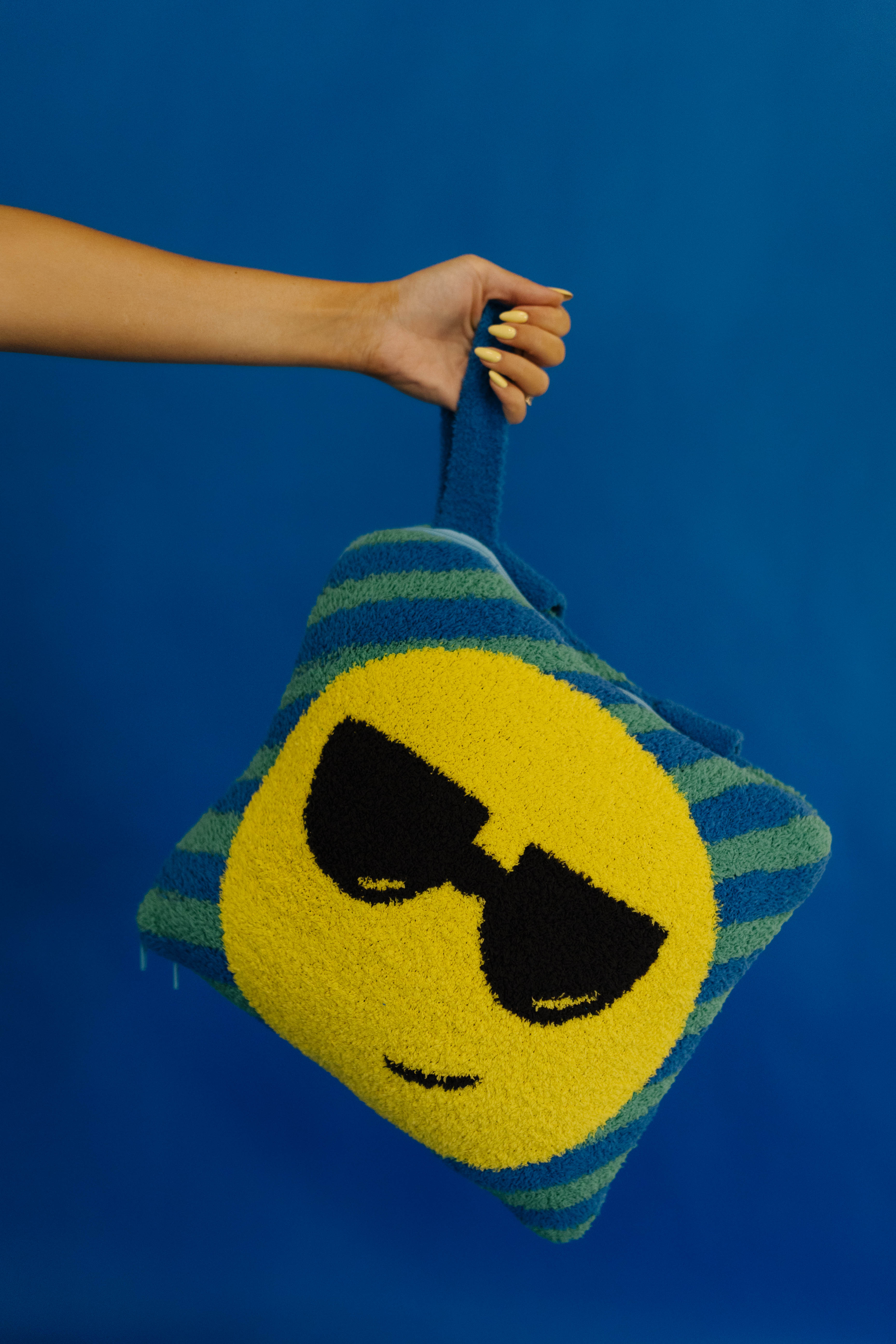 A hand holds a fuzzy quillow with black sunglasses and blue-green striped sides against a solid blue background.