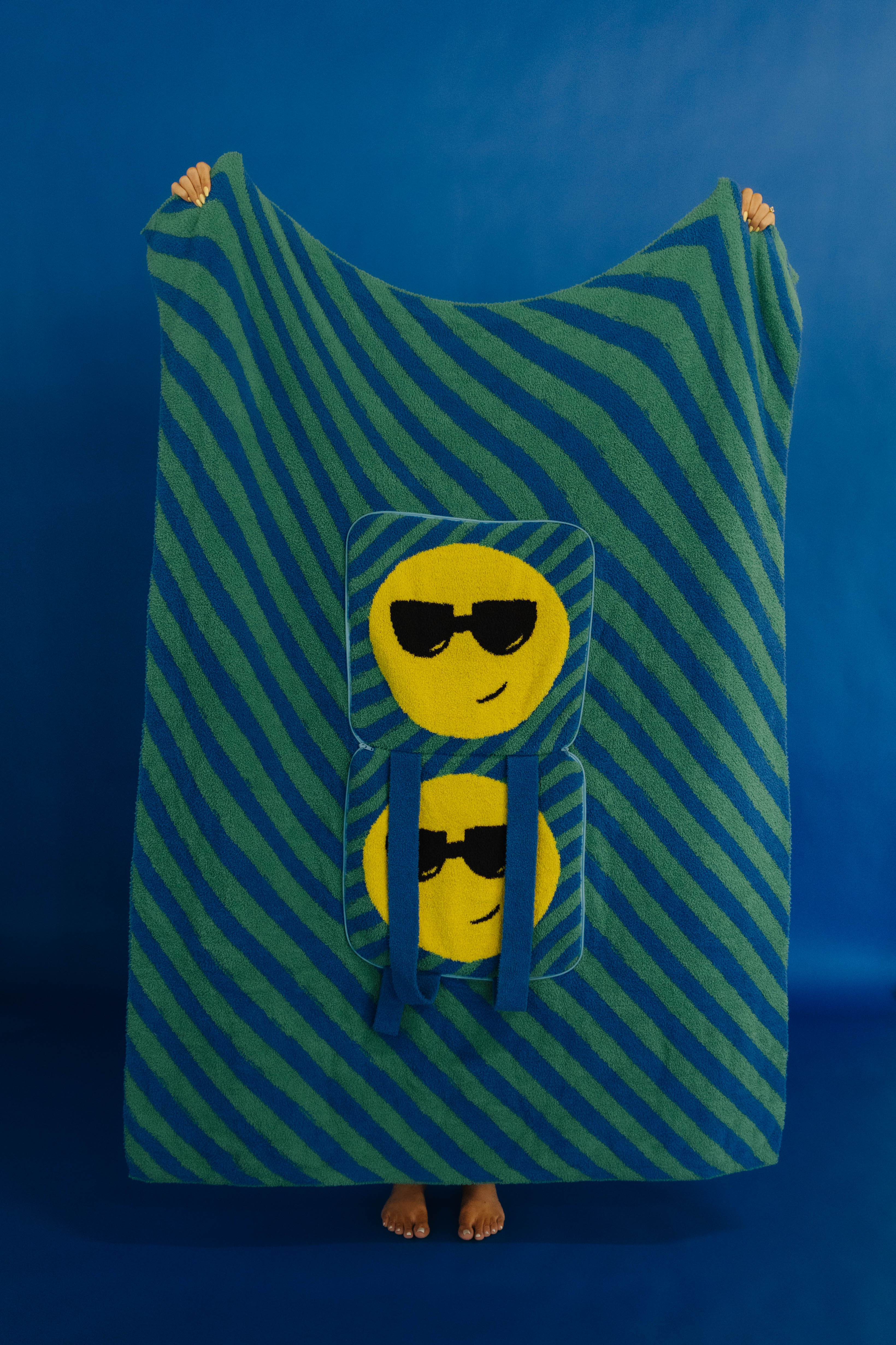 A person standing barefoot against a blue studio background, holding up a large striped quillow with two yellow smiley faces wearing sunglasses printed on the front.