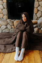 Model wrapped in the brown waffle blanket beside a stone fireplace.