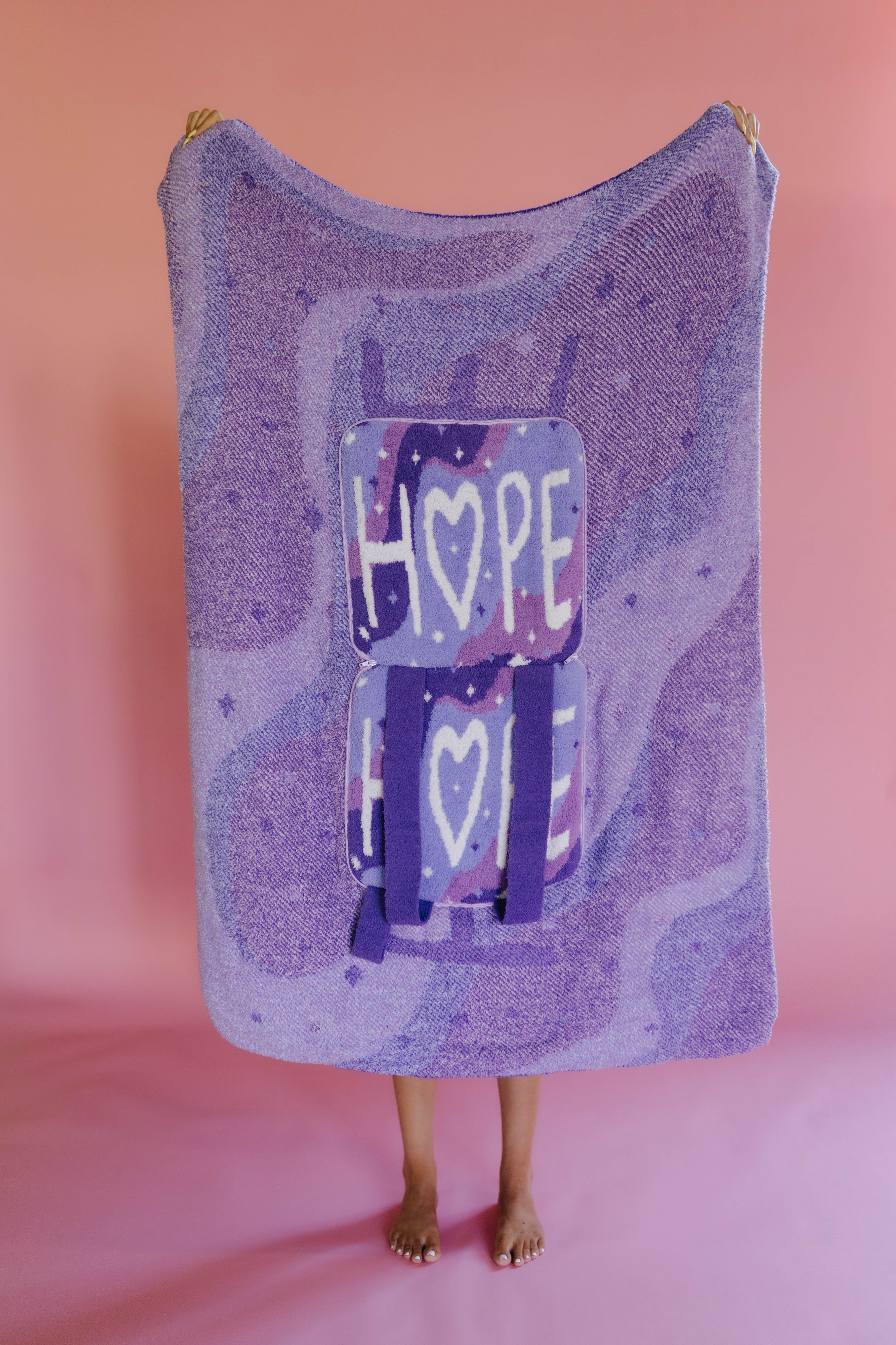 A person holding up a quillow, with backpack straps and a graphic featuring the word 'HOPE' and hearts, standing against a solid pink backdrop.