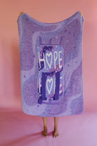 A person holding up a quillow, with backpack straps and a graphic featuring the word 'HOPE' and hearts, standing against a solid pink backdrop.
