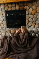 Two models sharing the brown waffle blanket while seated by a fireplace.
