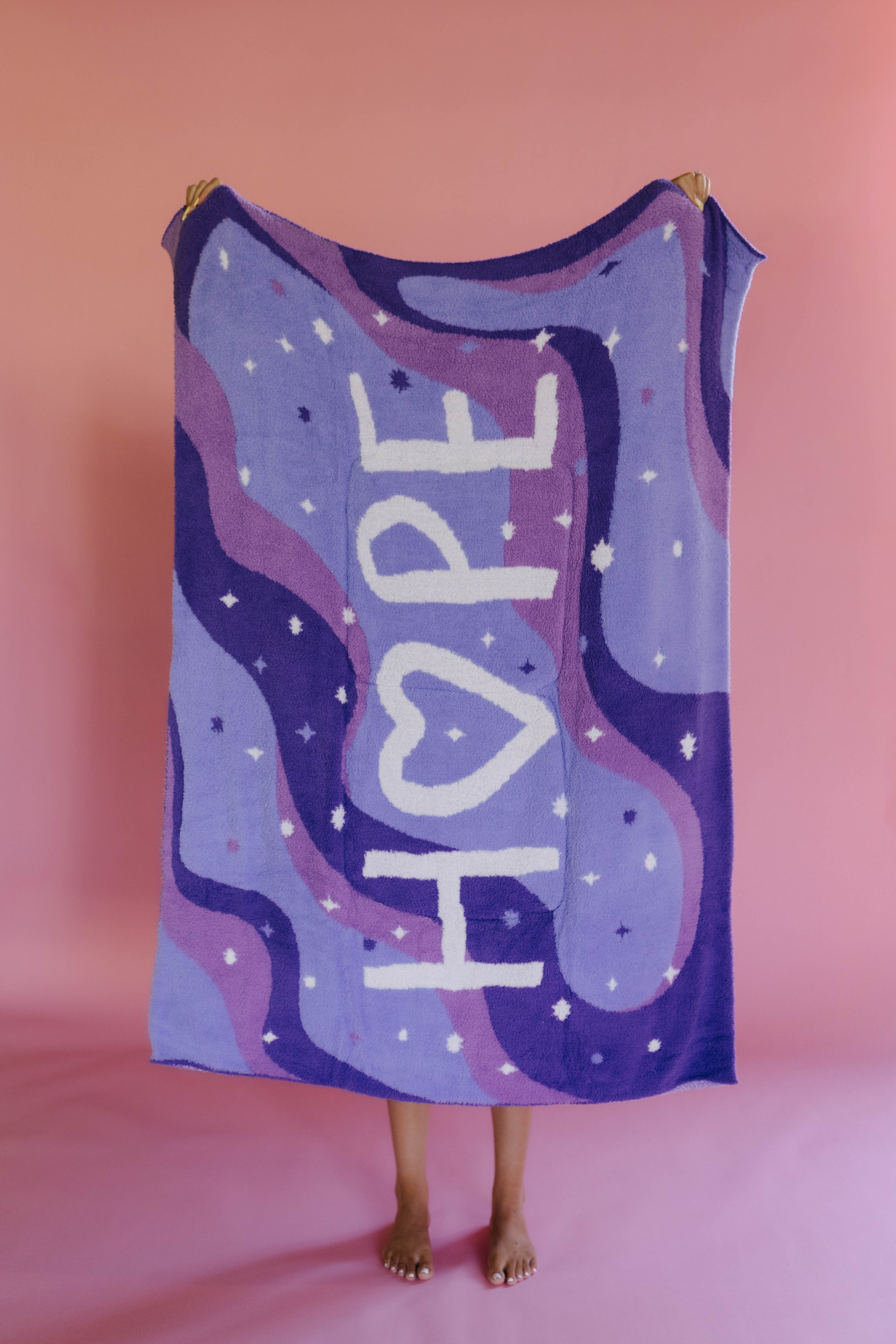 A person holds up a quillow in a purple and lavender wavy pattern, white stars, and the word 'HOPE' written vertically in white. The background is a solid pink studio backdrop.