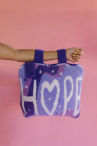 A person's arm and hand holding a purple and lavender quillow with the word 'HOPE' knitted in white, set against a solid pink background.