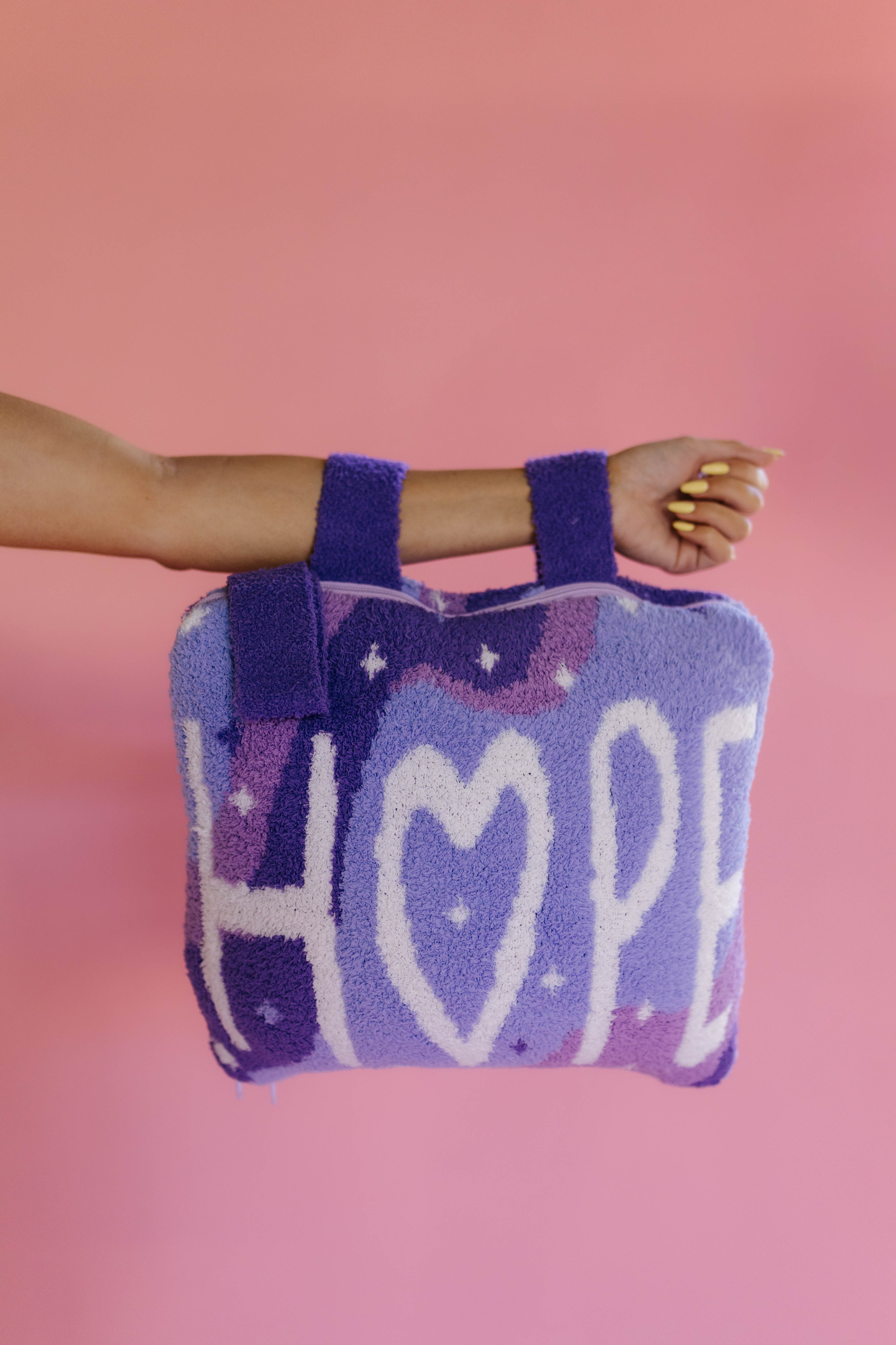 A person's arm and hand holding a purple and lavender quillow with the word 'HOPE' knitted in white, set against a solid pink background.