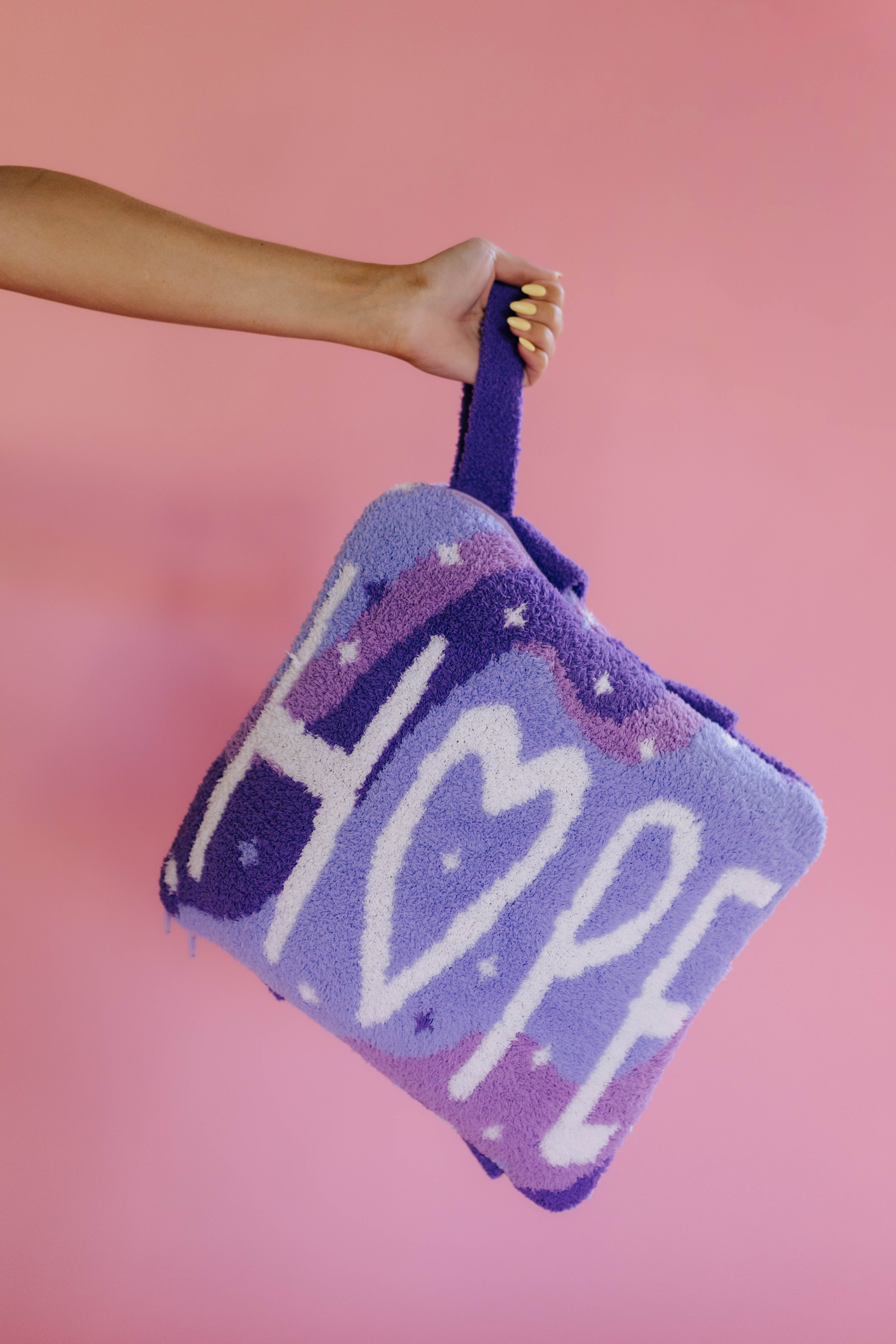 A hand holds a purple quillow with the word 'HOPE' in white letters, set against a solid pink background.