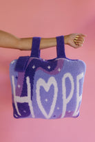 A fuzzy lavender quillow with the word 'HOPE' in white letters, held by a person's arm against a solid pink background.