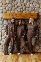 Three models wearing brown bow sweatshirts and sweatpants by a stone fireplace.