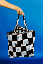Black and white checkered tote bag with sea-themed patterns held by a hand against a blue background