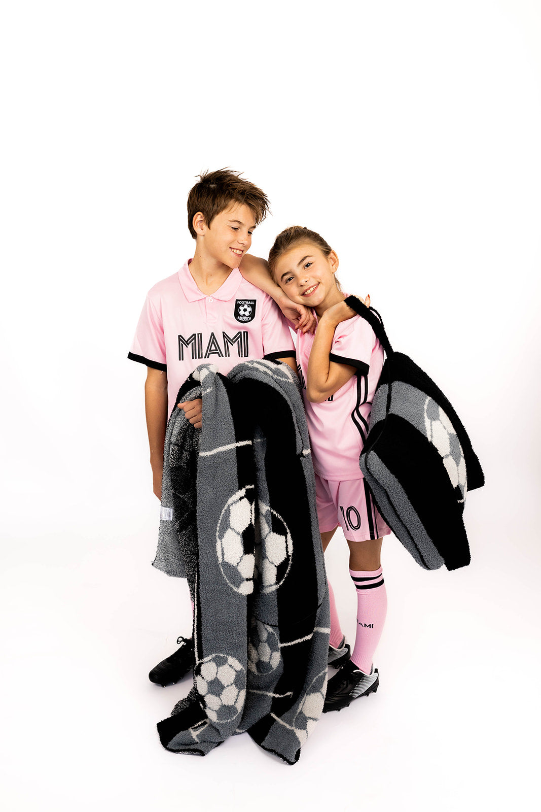 Two children in pink sports jerseys with 'MIAMI' text, holding a gray blanket with soccer ball patterns on a white background.