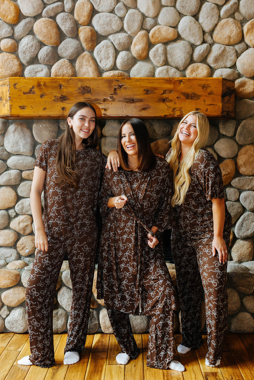 Three models in matching brown bow print pajamas, with two wearing matching robes.