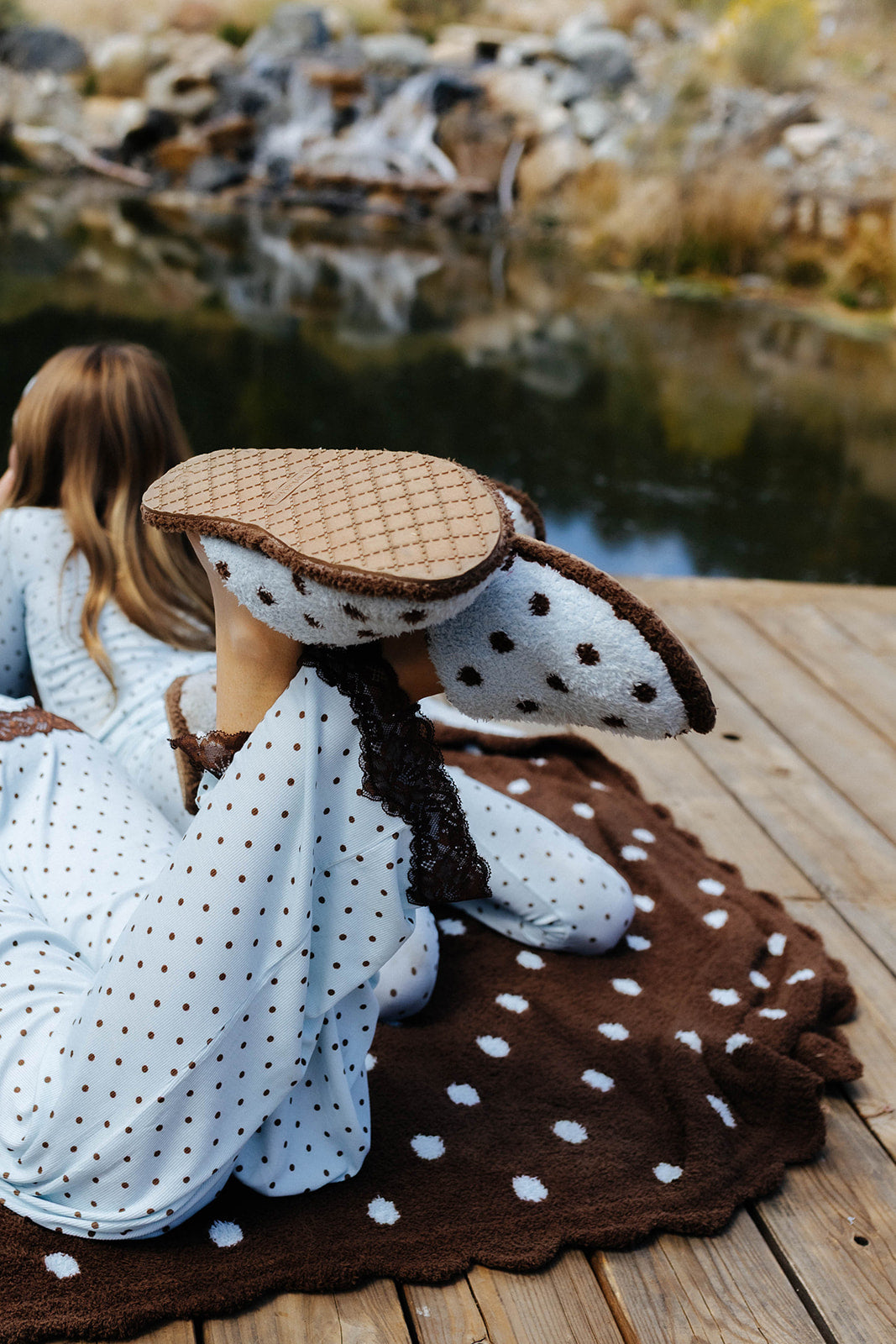 Polka dot slippers worn on a dock, with one lifted to show the brown textured sole.