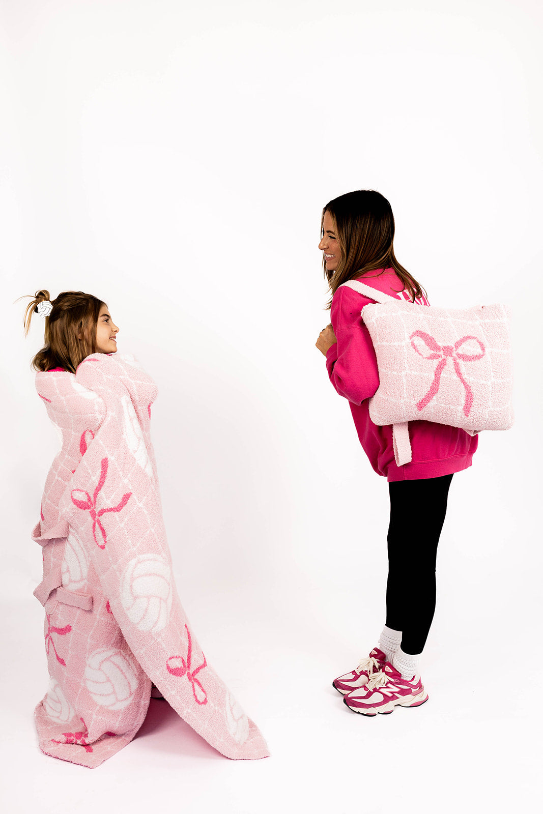 Two people, one wrapped in a pink volleyball blanket with bows and the other holding the quillow, on a white background.