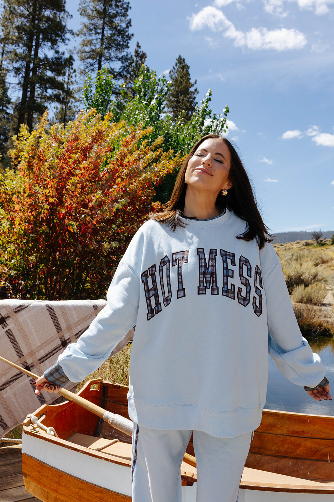 Model wearing the light blue Hot Mess sweatshirt and sweatpants beside a wooden boat.