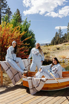 Three models in light blue Hot Mess sweatshirts and sweatpants beside a boat and plaid blanket.
