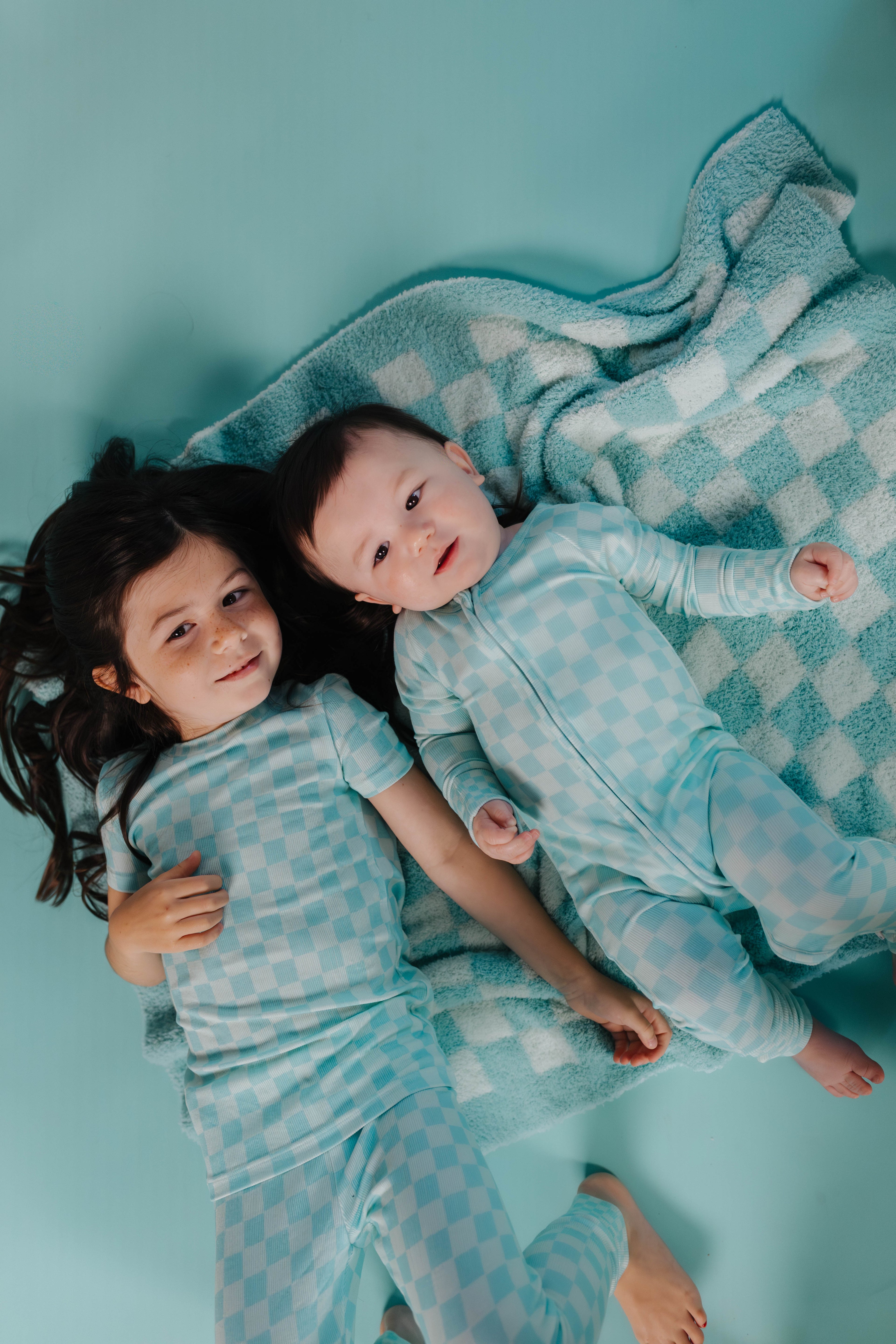 Two children in matching pajamas lying on a checkered blanket against a teal background