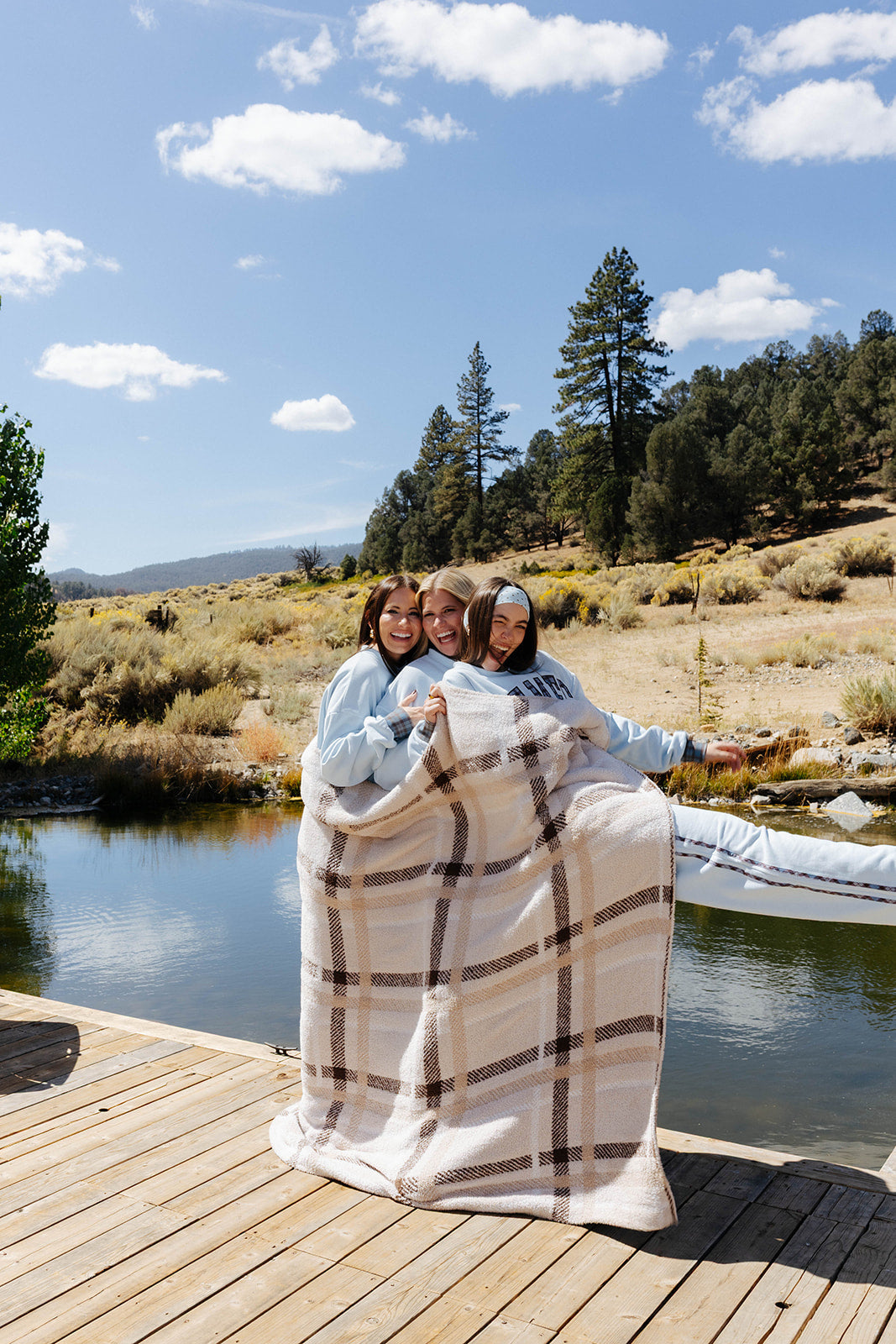 Three models wrapped together in a beige and brown plaid blanket on a lakeside dock.