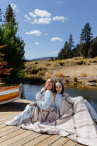 Three models seated together on a dock with the plaid blanket across their laps.