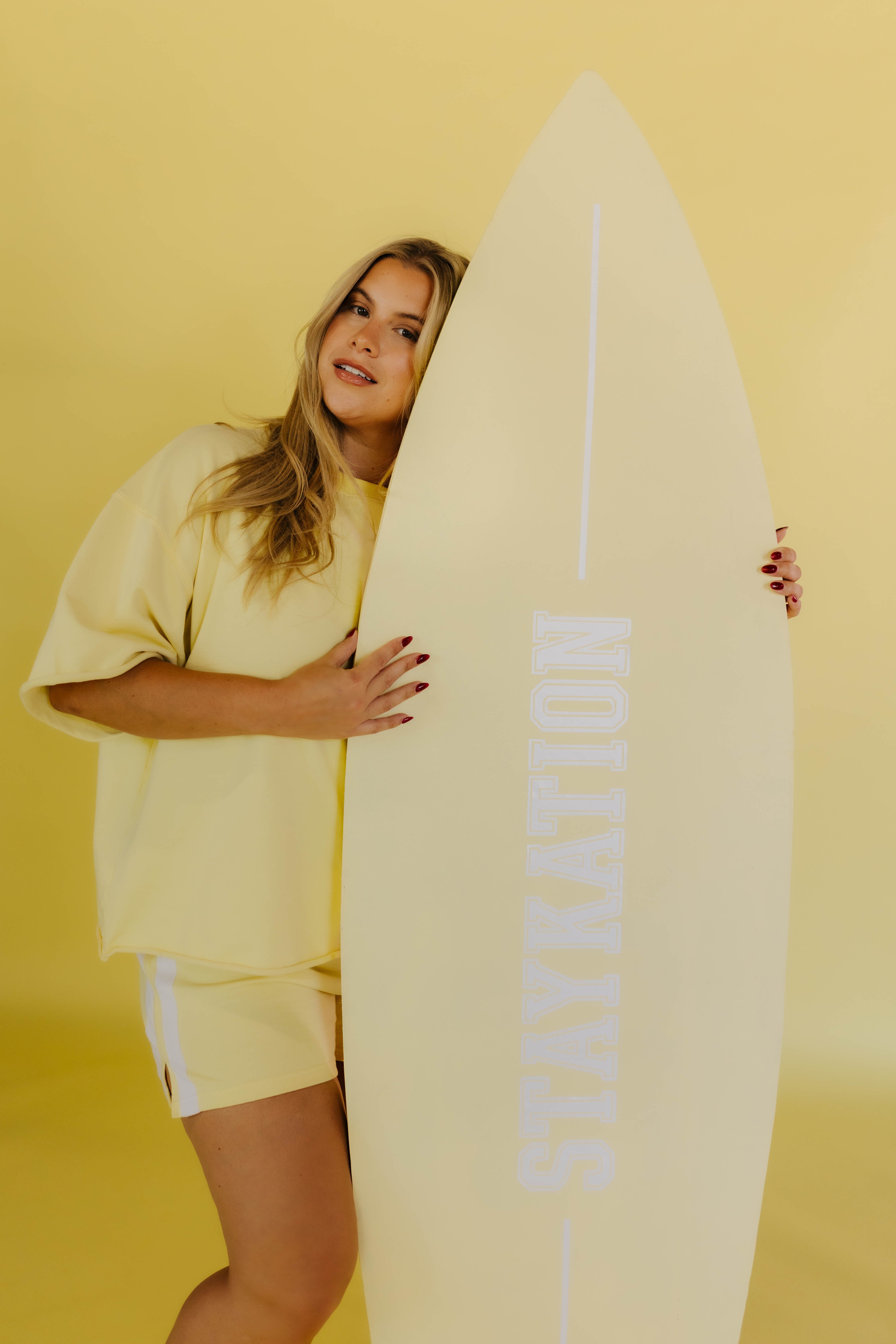 Woman holding a surfboard with 'STAYKATION' branding against a yellow background