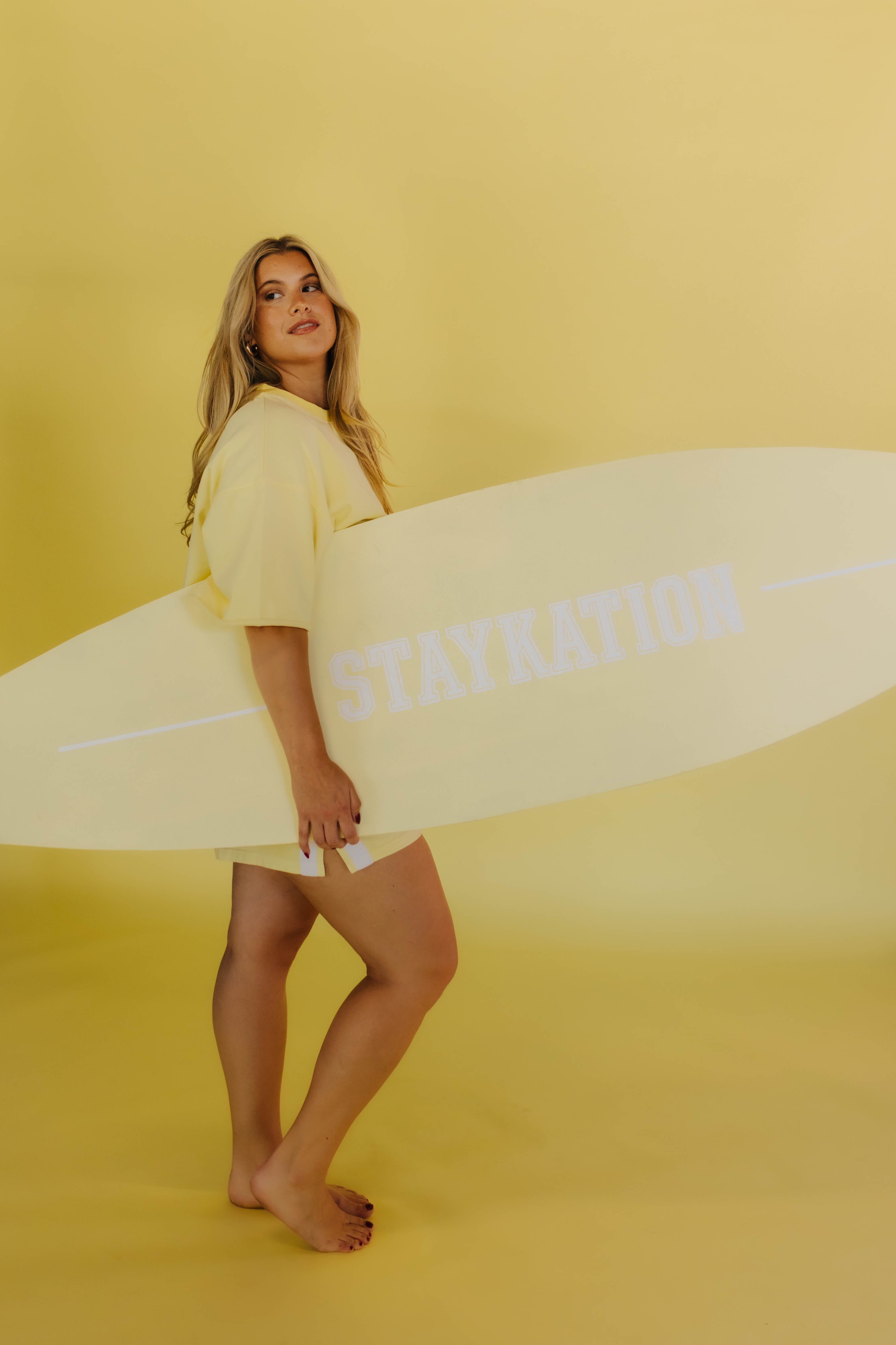 Woman holding a surfboard with 'STAYRATION' text on a yellow background