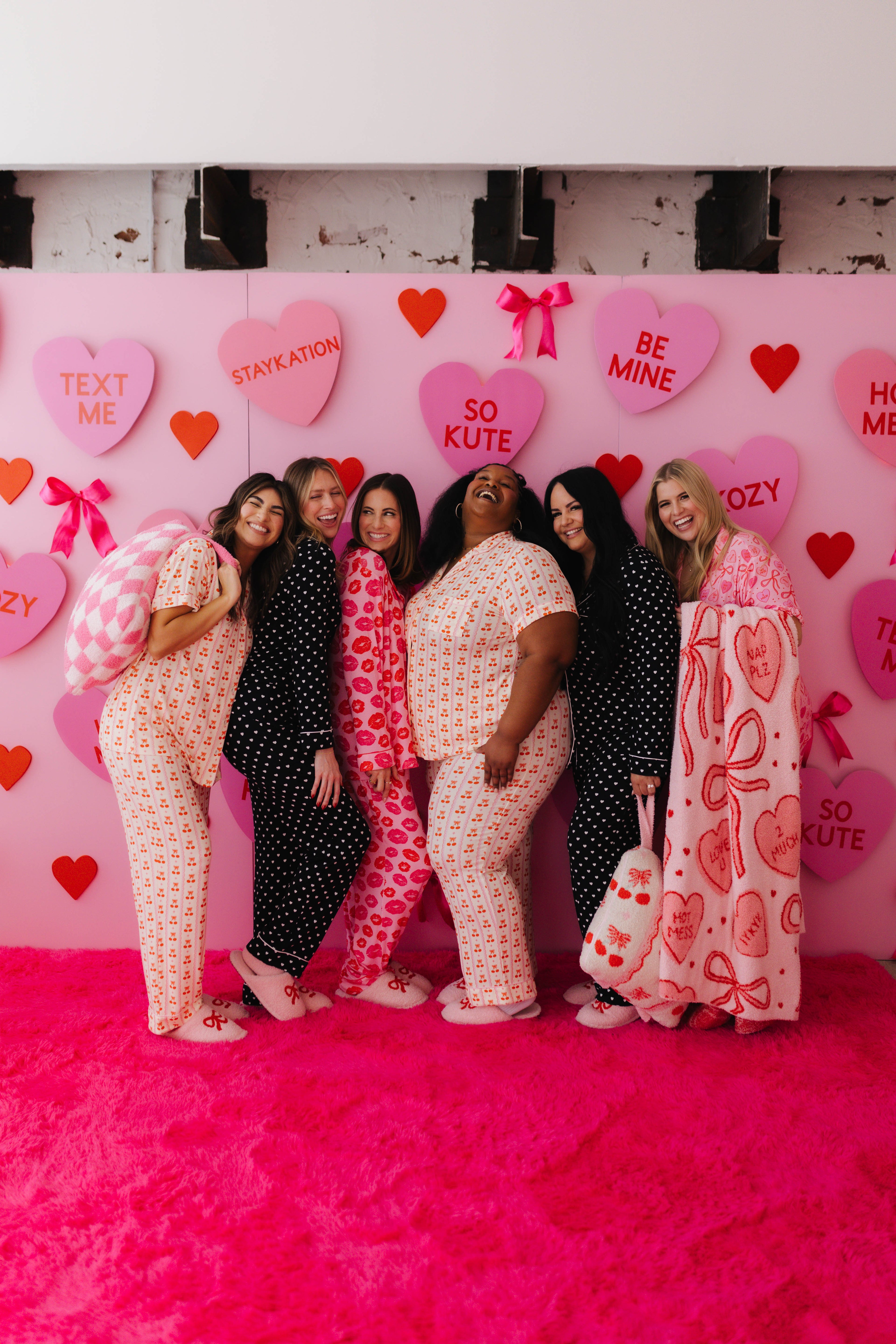 Group of women posing in front of a pink wall with heart-shaped decorations and text.