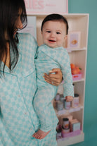 Woman holding a baby in a teal check outfit in a room with shelves in the background.