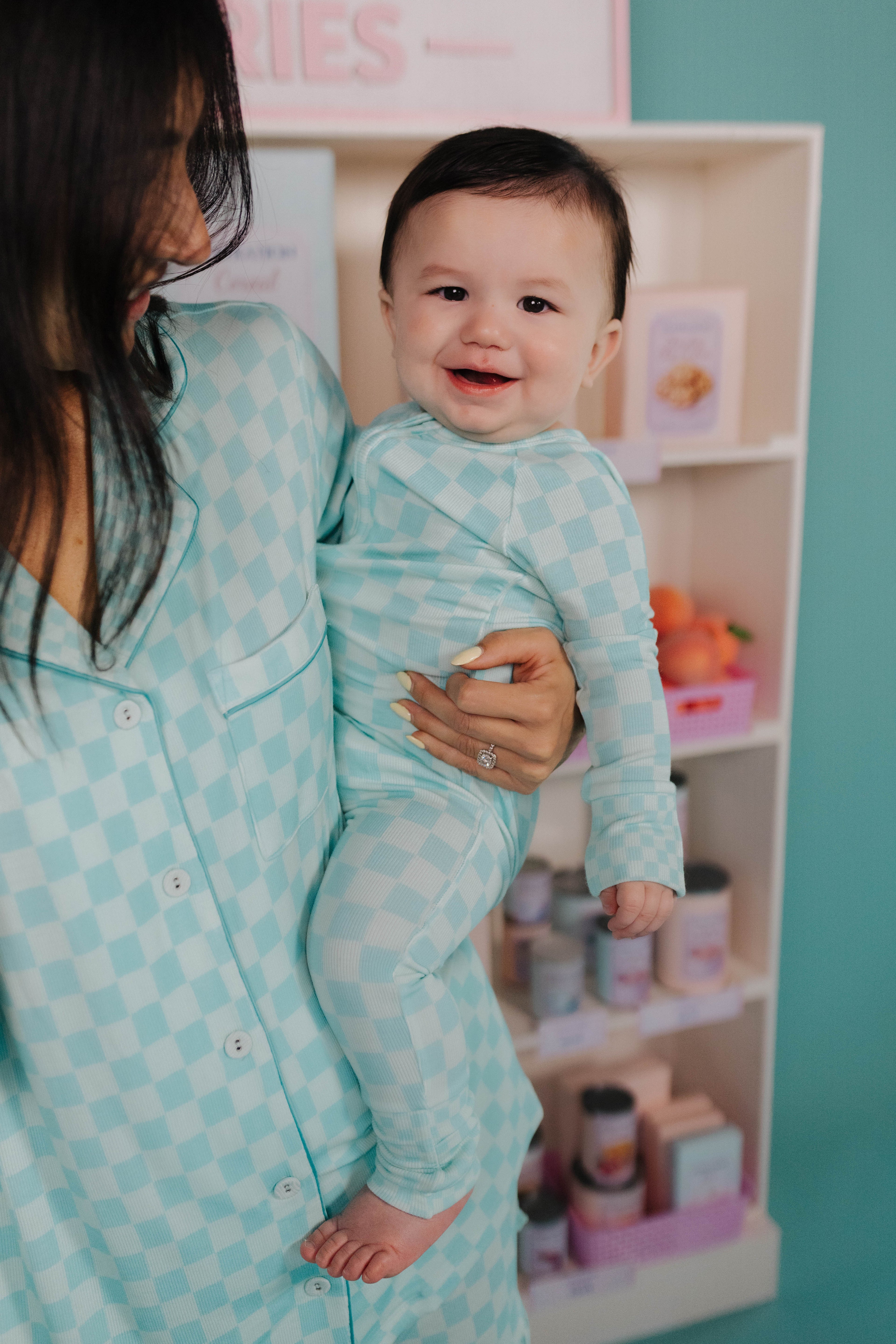 Woman holding a baby in a teal check outfit in a room with shelves in the background.