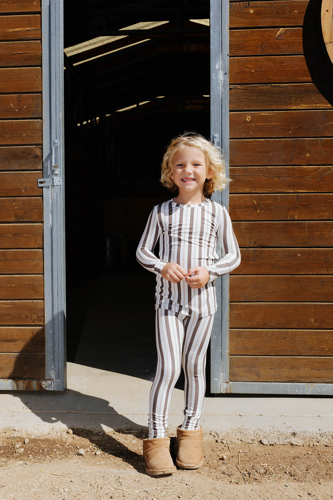Child smiling in the brown stripe pajama set at a stable doorway.