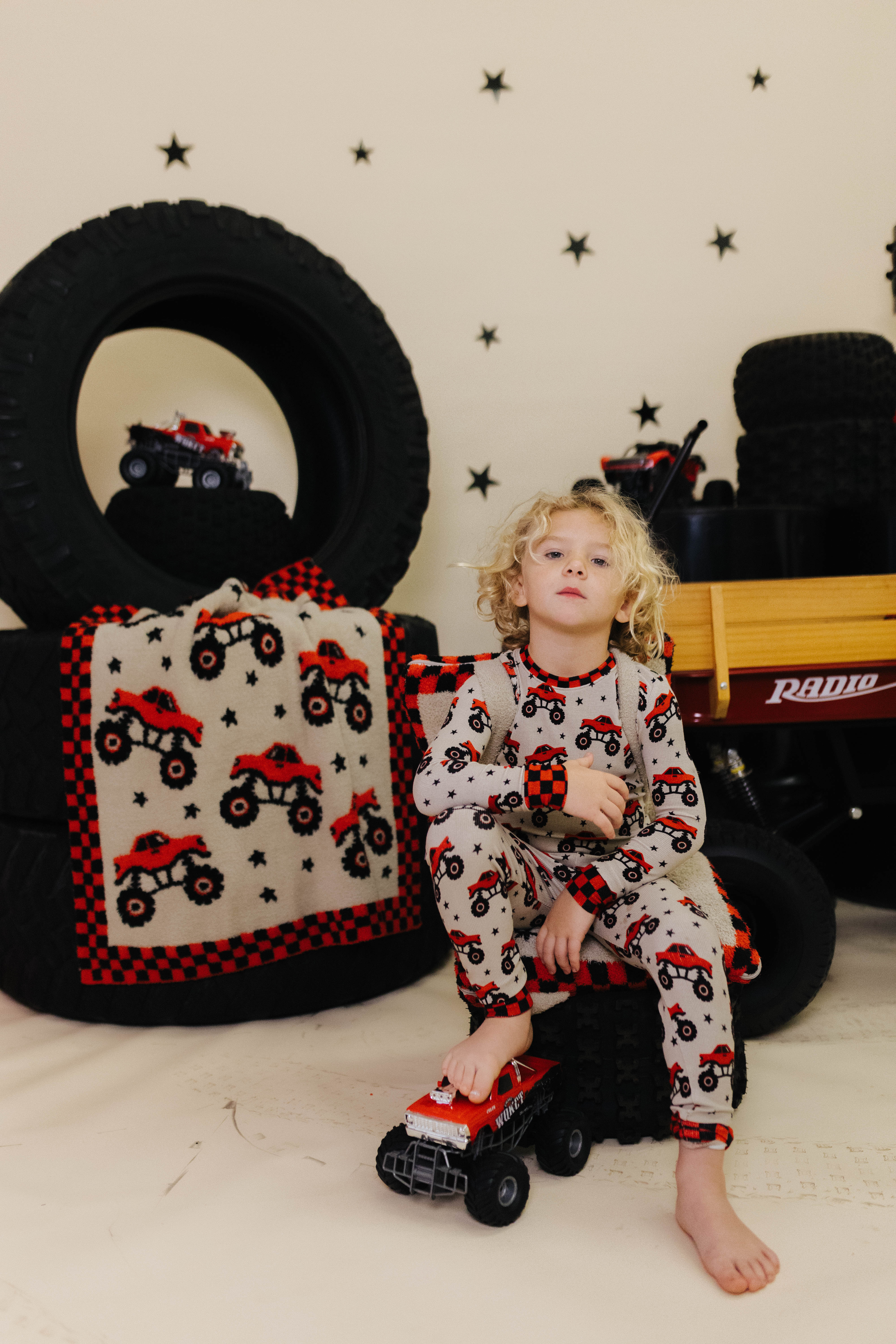 Child in pajamas with truck pattern sitting on a toy truck surrounded by tires and a blanket with truck design.