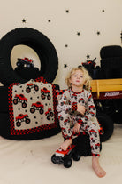 Child in pajamas with truck pattern sitting on a toy truck surrounded by tires and a blanket with truck design.