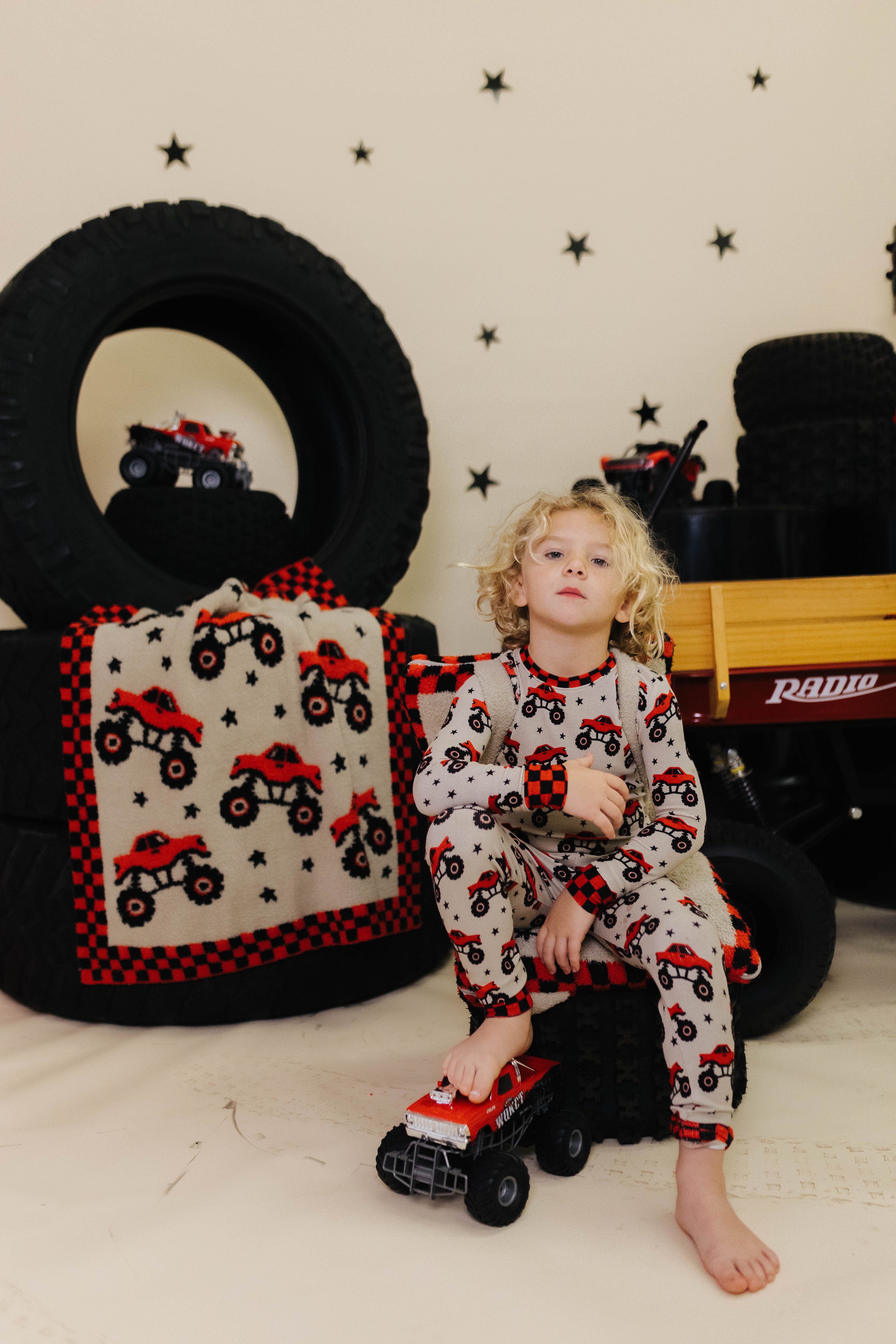 Child in pajamas with truck pattern sitting on a toy truck surrounded by tires and a blanket with truck design.