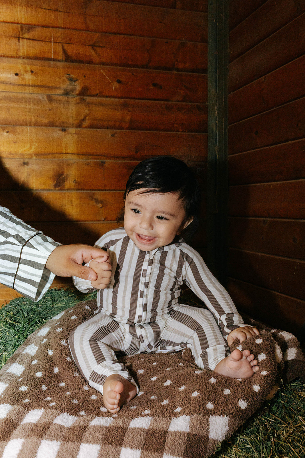 Baby smiling in brown stripe zip pajamas while held beside a hay bale.