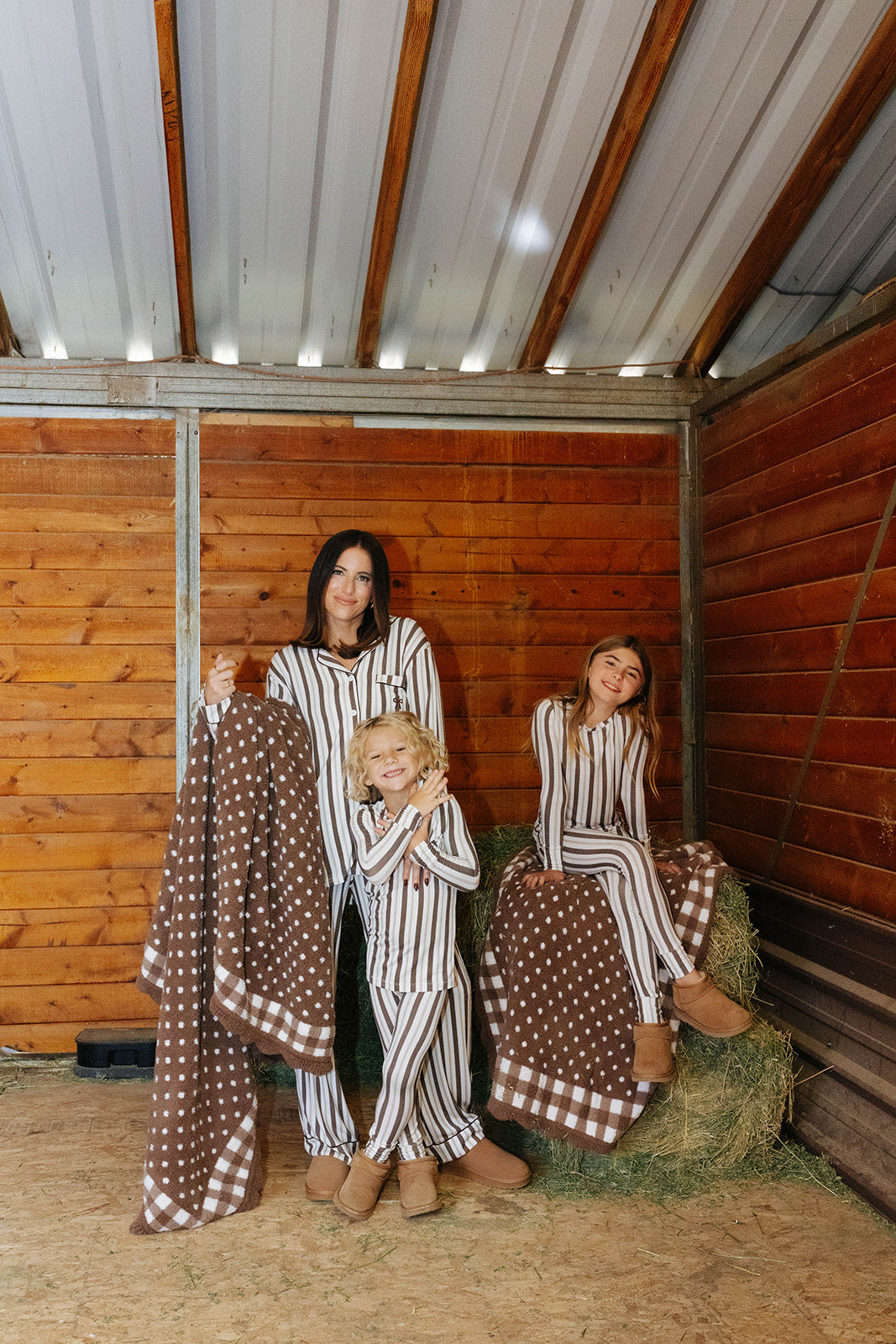 Adult and children wearing brown and cream striped pajamas and holding brown polka dot blankets in a wooden stable.