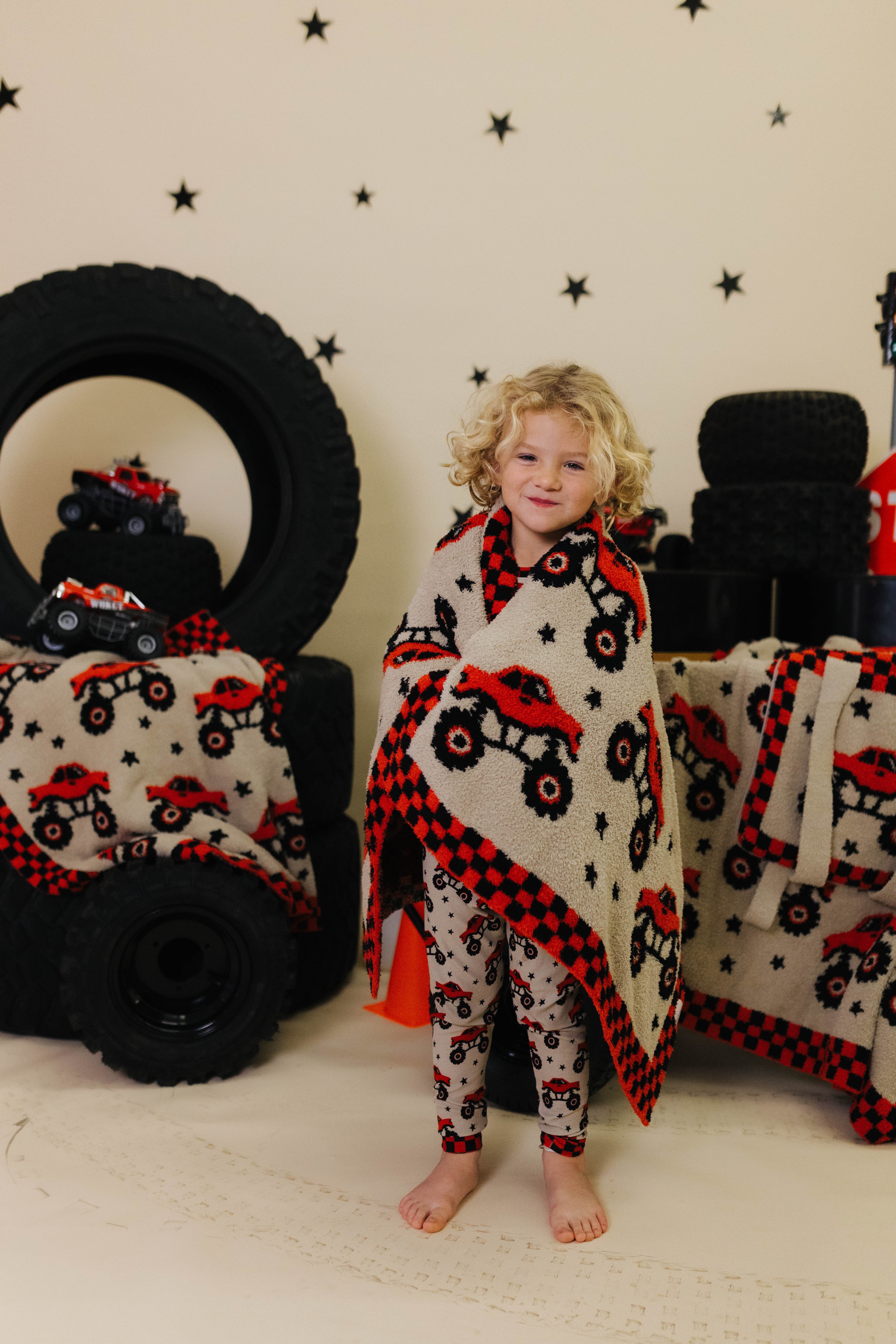 Child wrapped in a blanket with red and black patterns, standing in front of large tires and star-patterned wall.