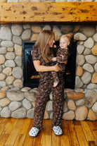 Adult holding a baby while wearing matching brown bow print pajamas by a fireplace.