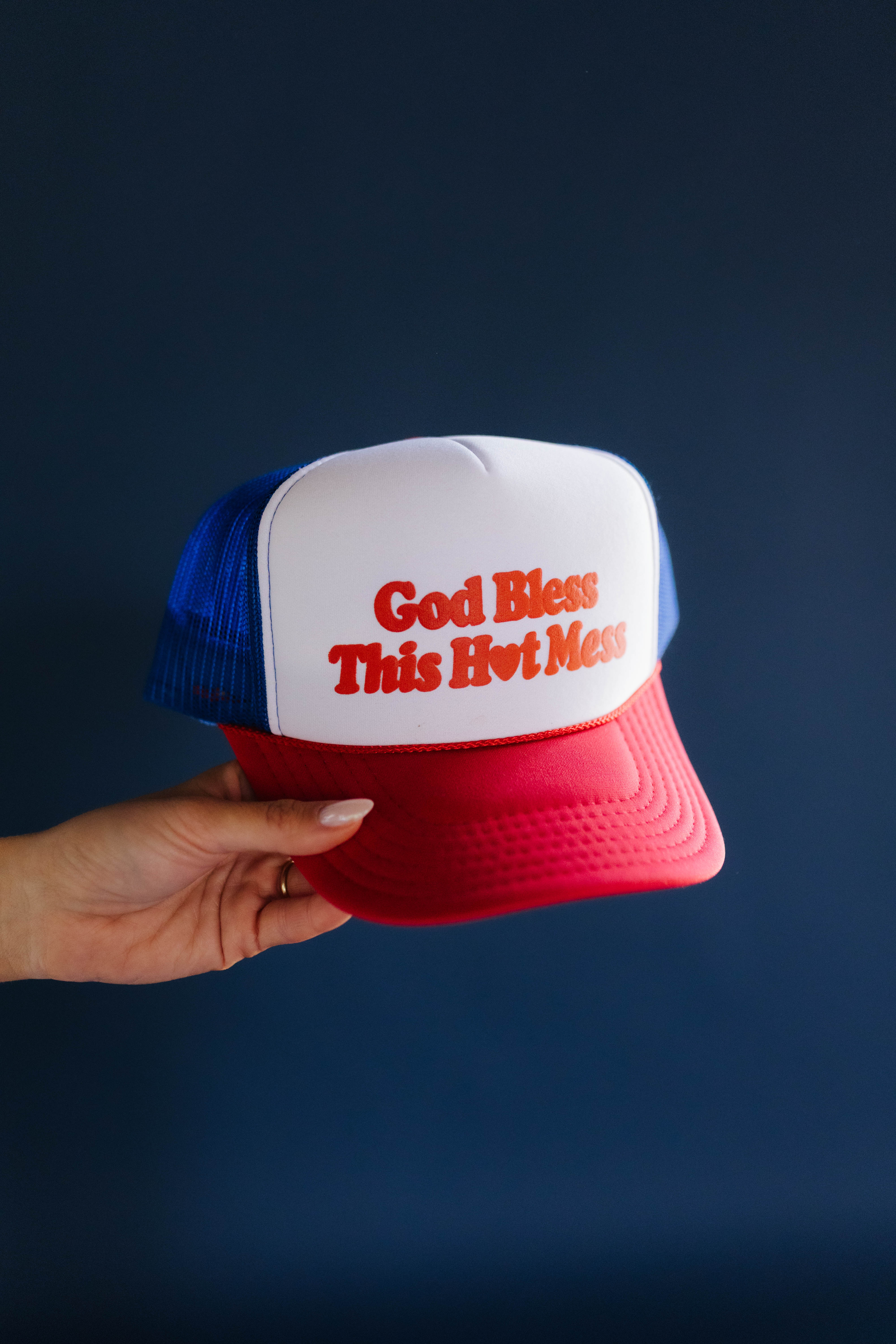 Trucker hat with 'God Bless This Hvit Moss' text held by a hand against a dark background