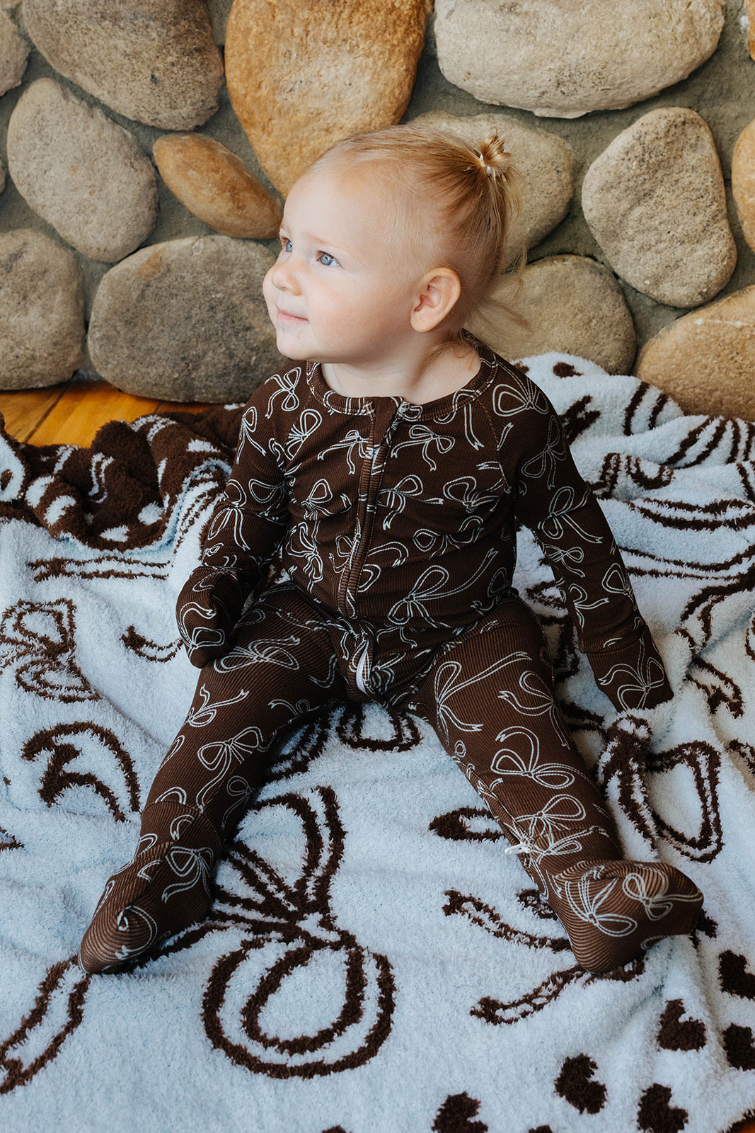 Side view of a baby wearing brown bow print zip pajamas on a matching bow quillow.