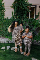 Adult holding a baby beside a child, all wearing matching brown check pajamas.