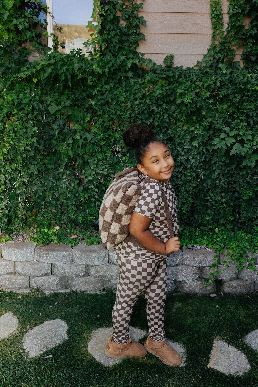 Side and back view of a child wearing brown check pajamas and a matching quillow backpack.