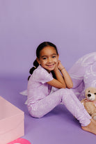 Young girl in a purple outfit sitting on a purple floor with a teddy bear.