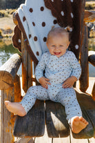 Baby smiling in blue and brown polka dot zip pajamas while seated in a wooden chair.