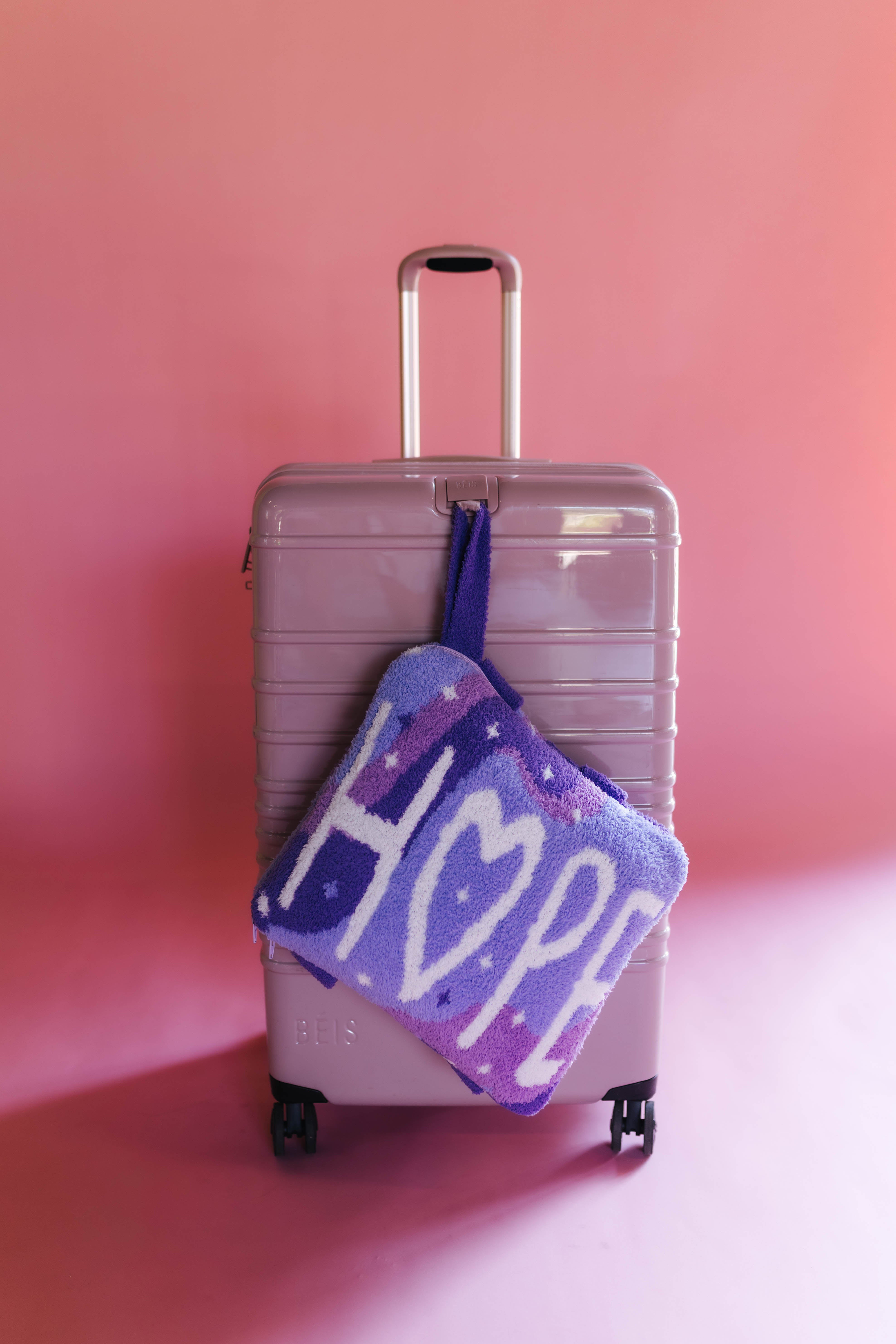 A lavender hard-shell suitcase standing against a pink studio background with the quillow hanging off of the front