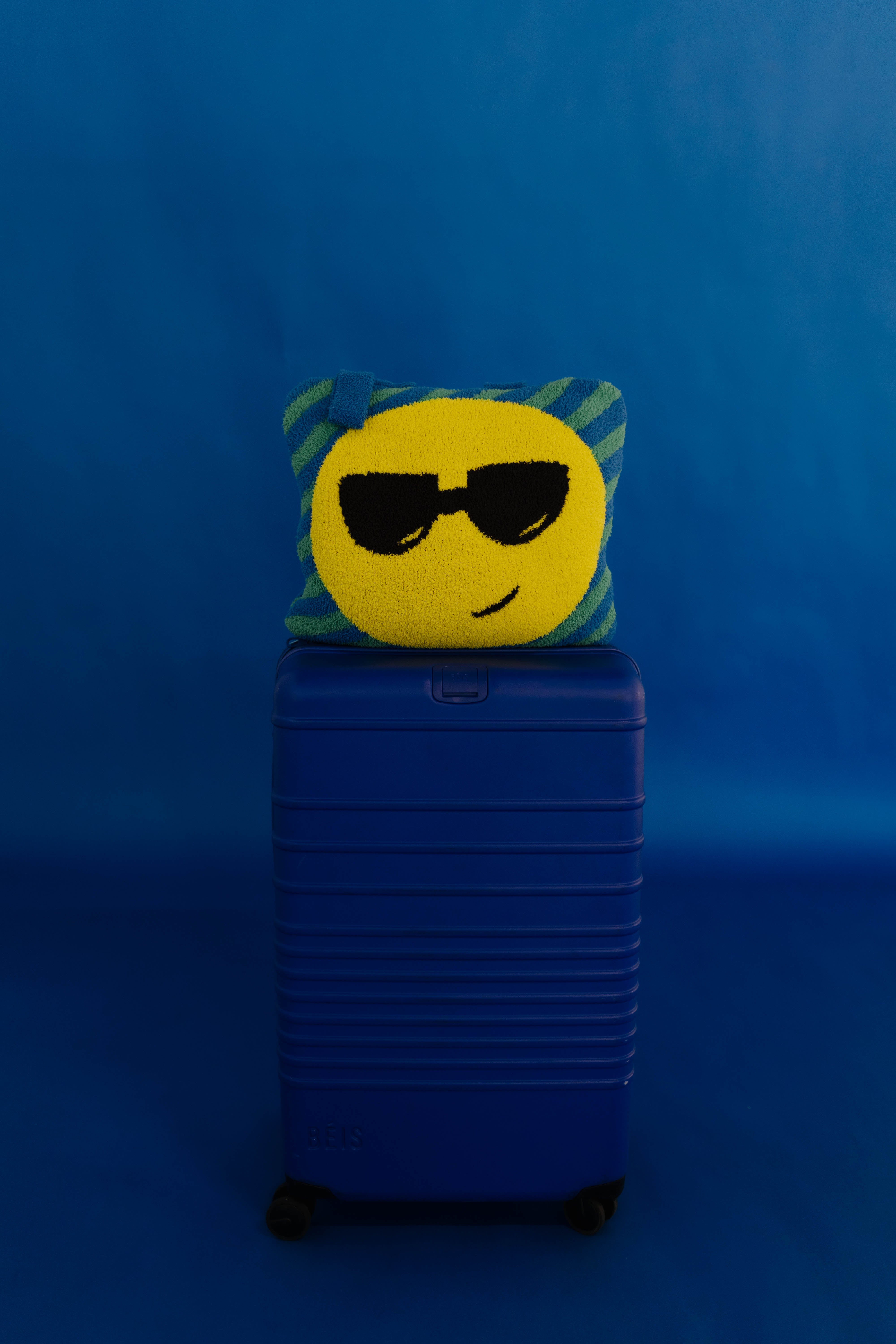 A yellow circular emoji face quillow with black sunglasses and a green striped border, resting on top of a blue hard-shell suitcase against a solid blue background.