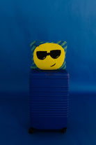 A yellow circular emoji face quillow with black sunglasses and a green striped border, resting on top of a blue hard-shell suitcase against a solid blue background.