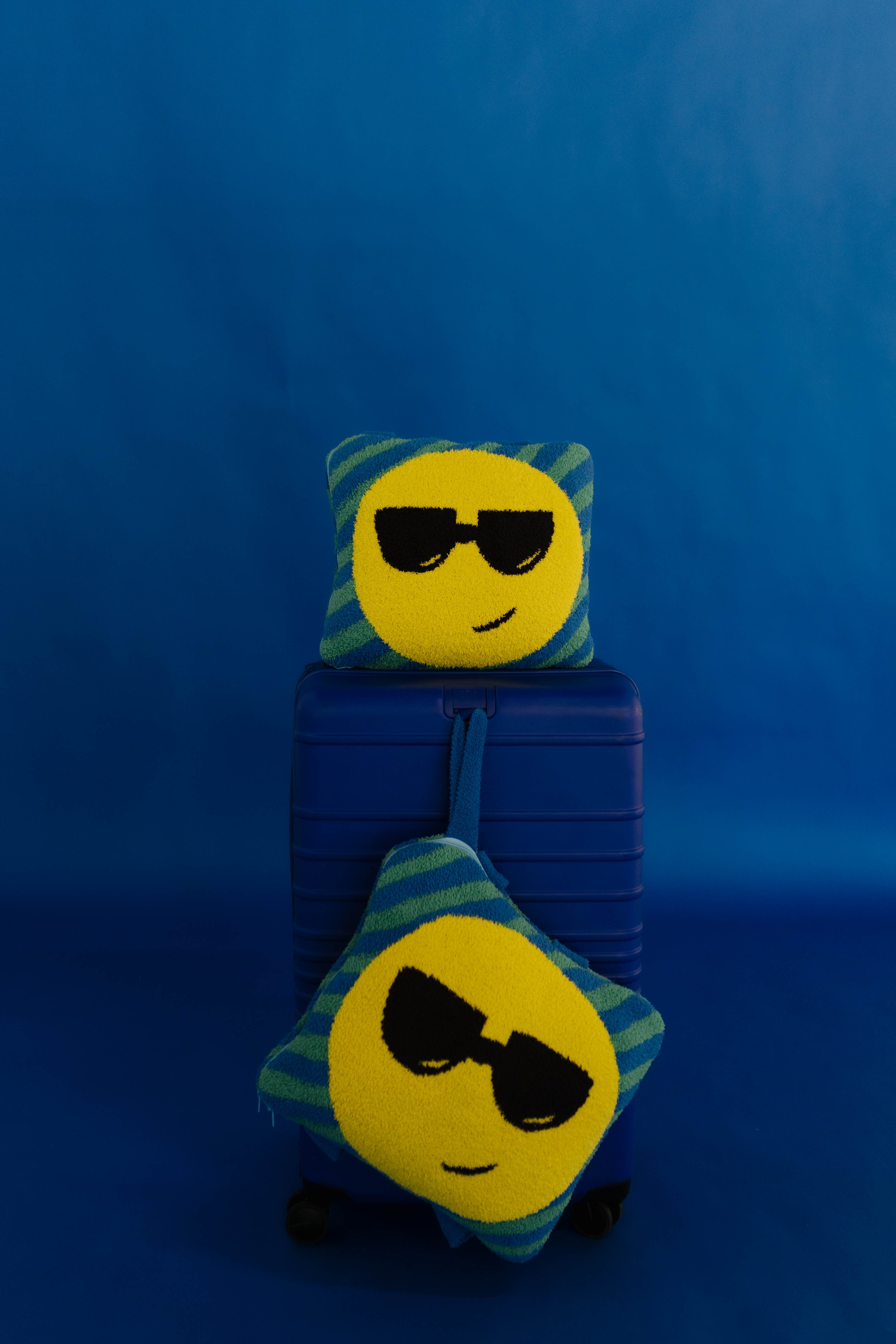 Two emoji face quillows with yellow circular faces wearing black sunglasses and blue-green striped edges, displayed on and in front of a blue suitcase against a blue background.