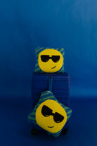 Two emoji face quillows with yellow circular faces wearing black sunglasses and blue-green striped edges, displayed on and in front of a blue suitcase against a blue background.